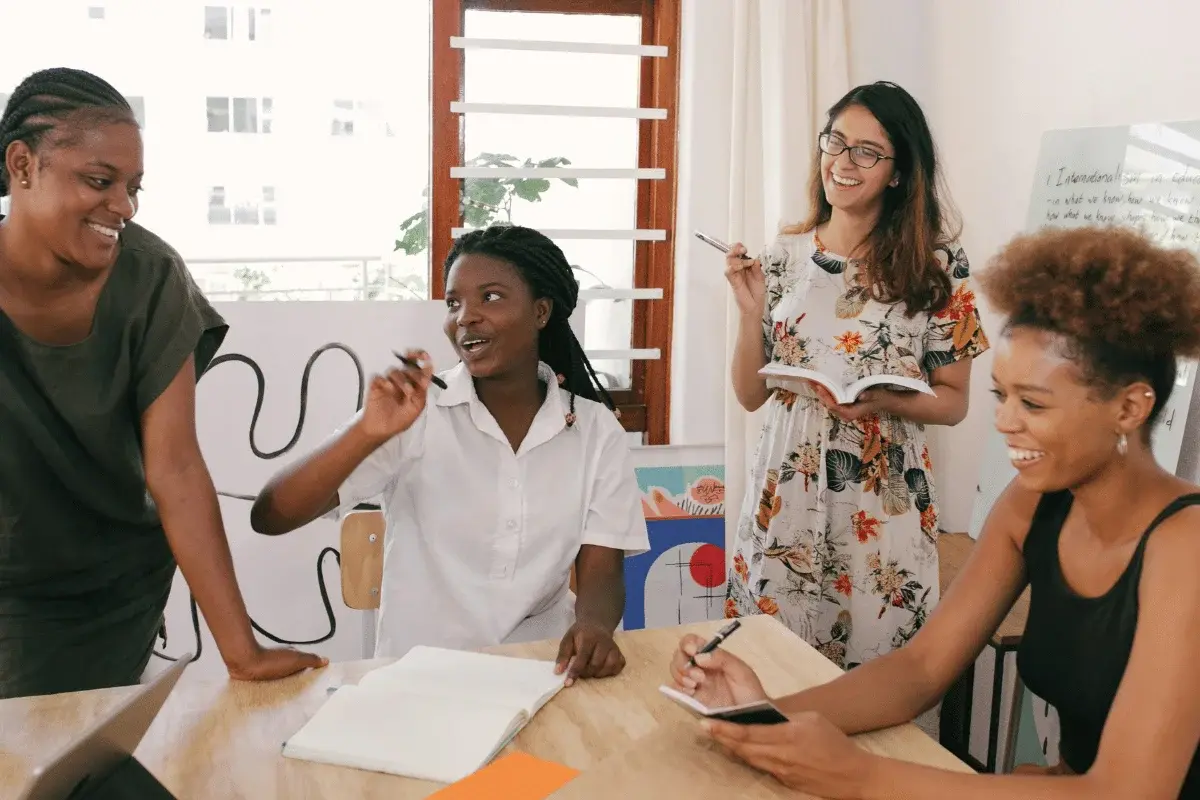 Four women collaborate enthusiastically around a table, surrounded by notes and a whiteboard, creating a lively and creative atmosphere.