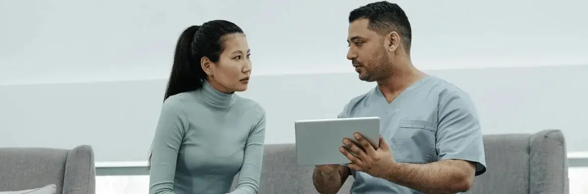 A healthcare professional in scrubs discusses information on a tablet with a seated patient in a modern office setting.