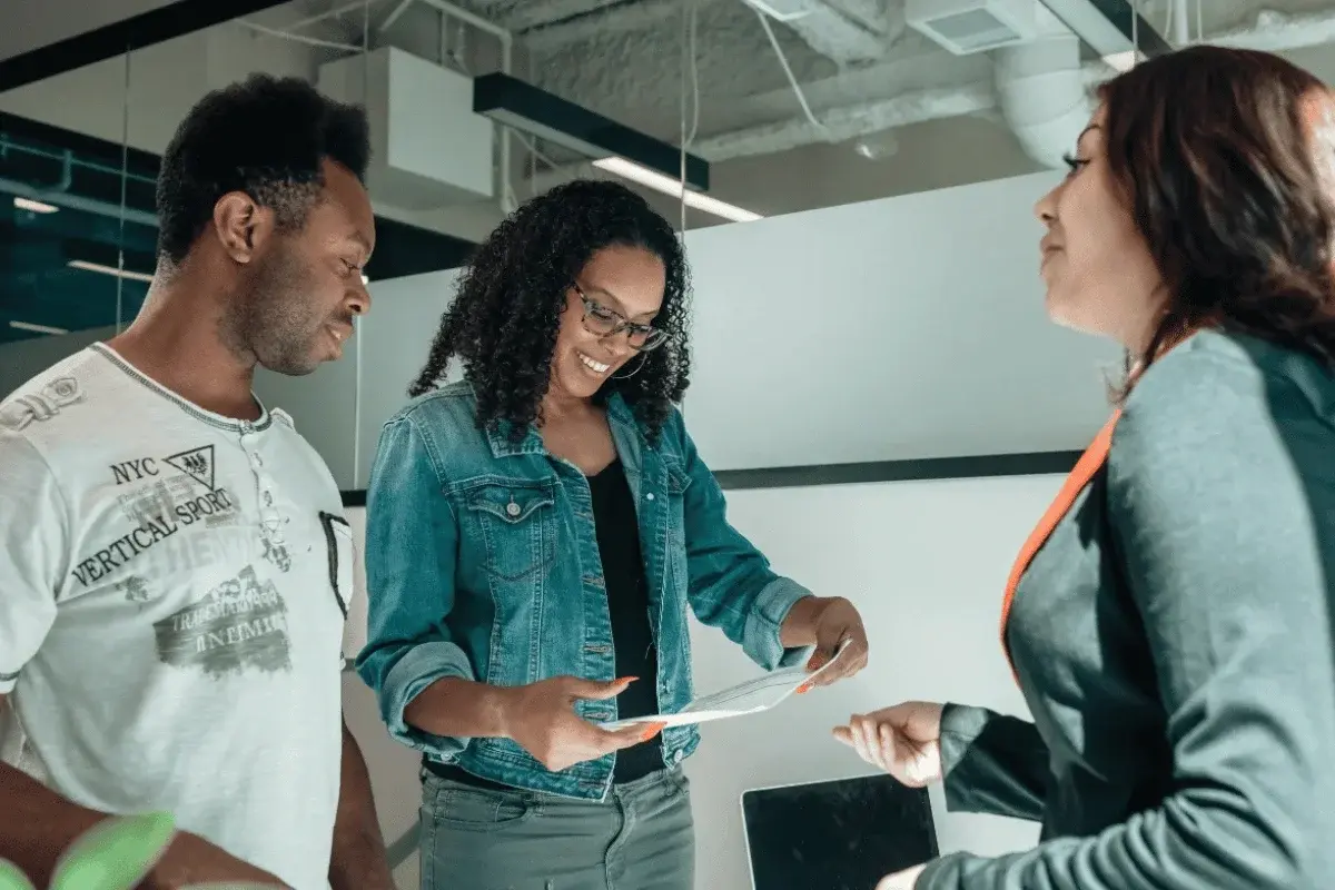 Three people in a modern office setting share a document. A woman in a denim jacket smiles, creating a collaborative and positive atmosphere.