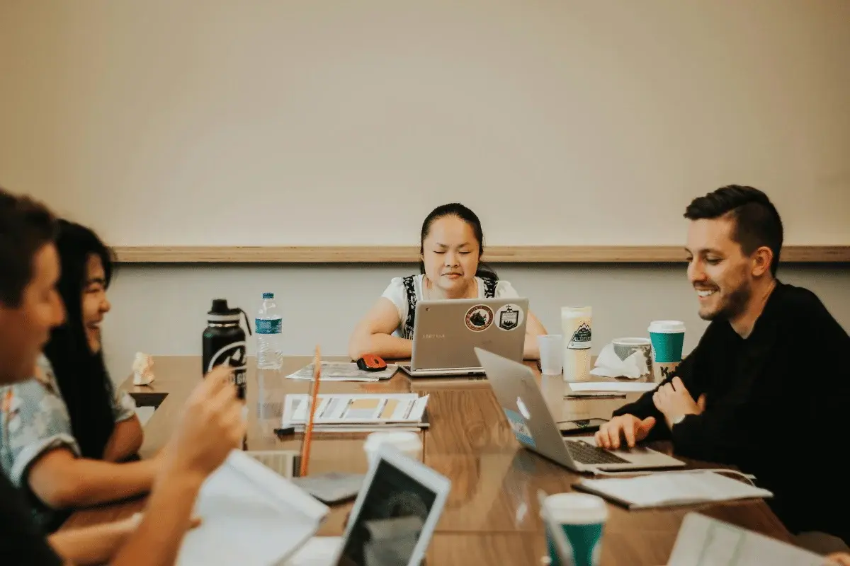 A group of people sit around a table engaged in discussion, with laptops, papers, and drinks visible. The atmosphere is collaborative and focused.