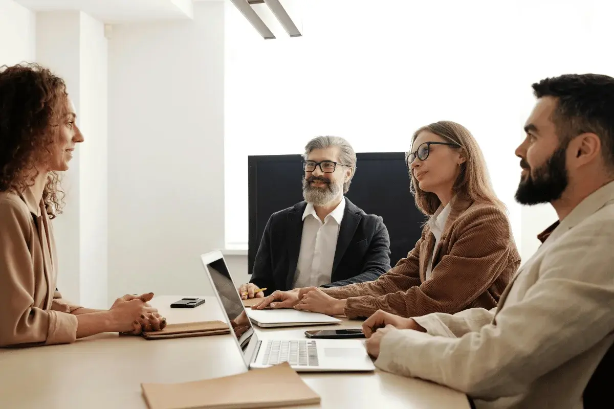 Four professionals engaged in a business meeting discussing hiring decisions around a conference table with laptops and notebooks.