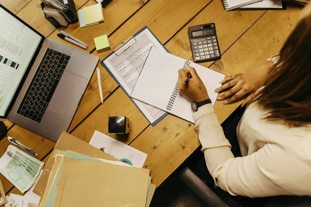 Overhead view of a person writing in a notebook on a wooden desk with a laptop, calculator, papers, and sticky notes, conveying focus and productivity.