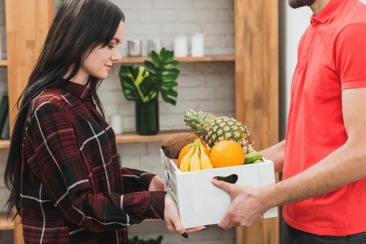 A person receives a crate filled with fresh fruits, including bananas, oranges, and a pineapple, in a cozy interior setting.