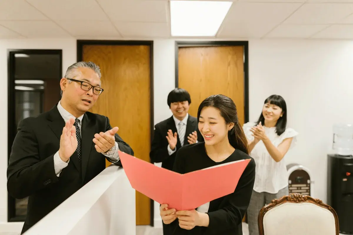 A woman holding a pink folder is smiling while three colleagues applaud her in an office setting. The atmosphere is positive and congratulatory.