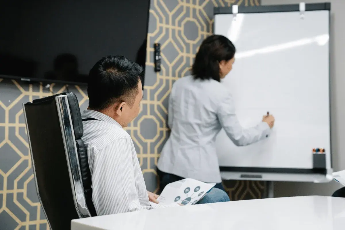 Businesswoman writing on a whiteboard while colleague observes during a meeting.