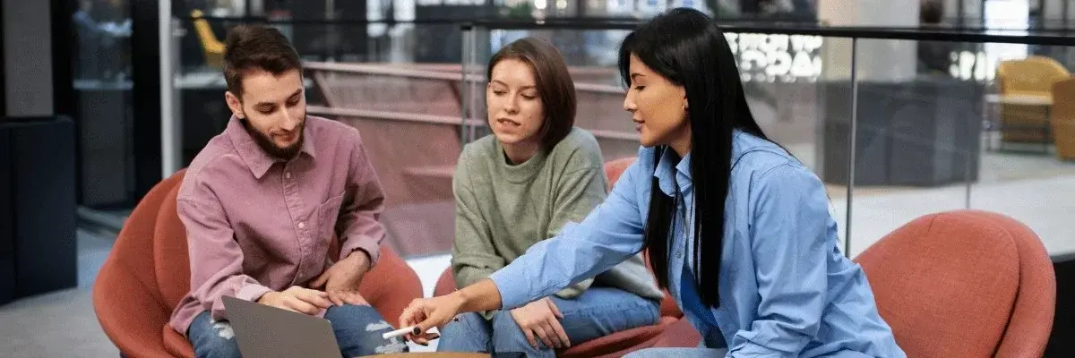Three individuals engage in a discussion around a table, featuring laptops and devices, in a modern indoor setting.