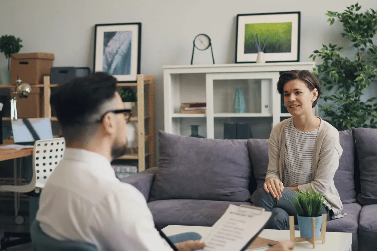 A man and a woman sit across from each other in a modern office with plants and art. The man holds a clipboard, suggesting a relaxed meeting.