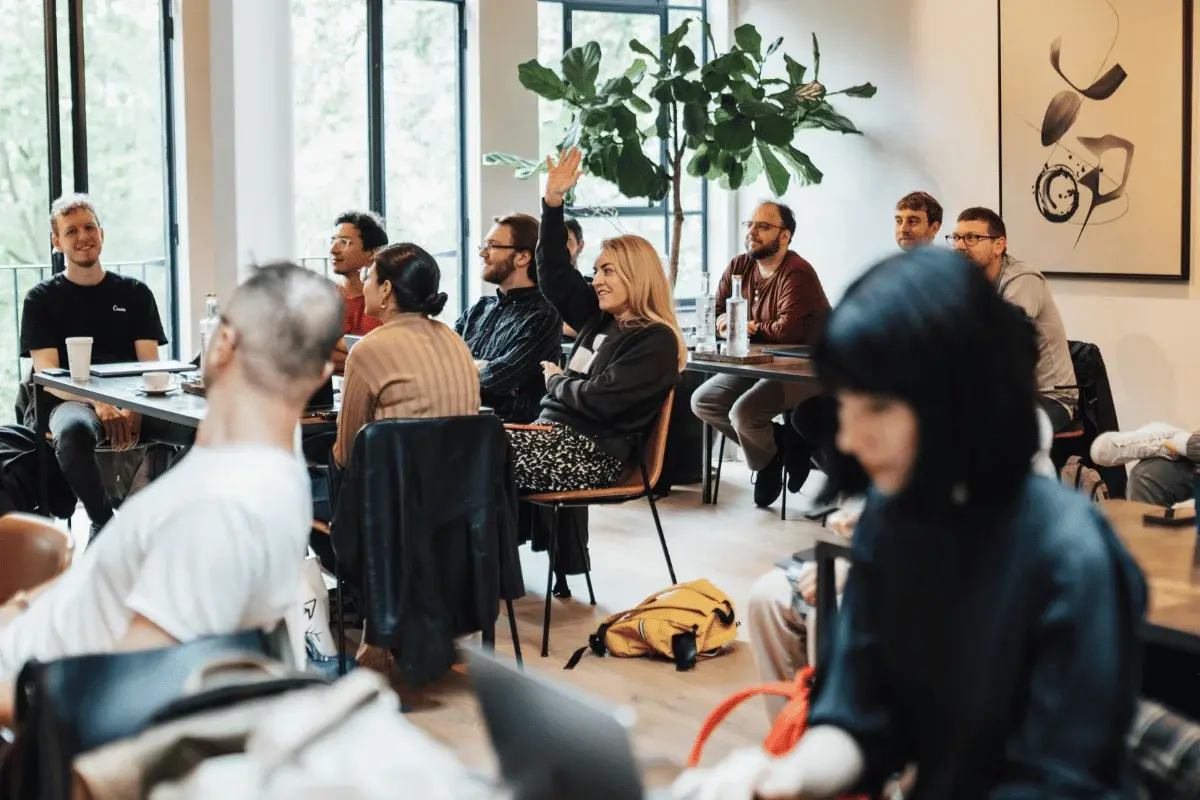 A group of people engaged in a collaborative discussion, sitting at tables in a well-lit room decorated with indoor plants.