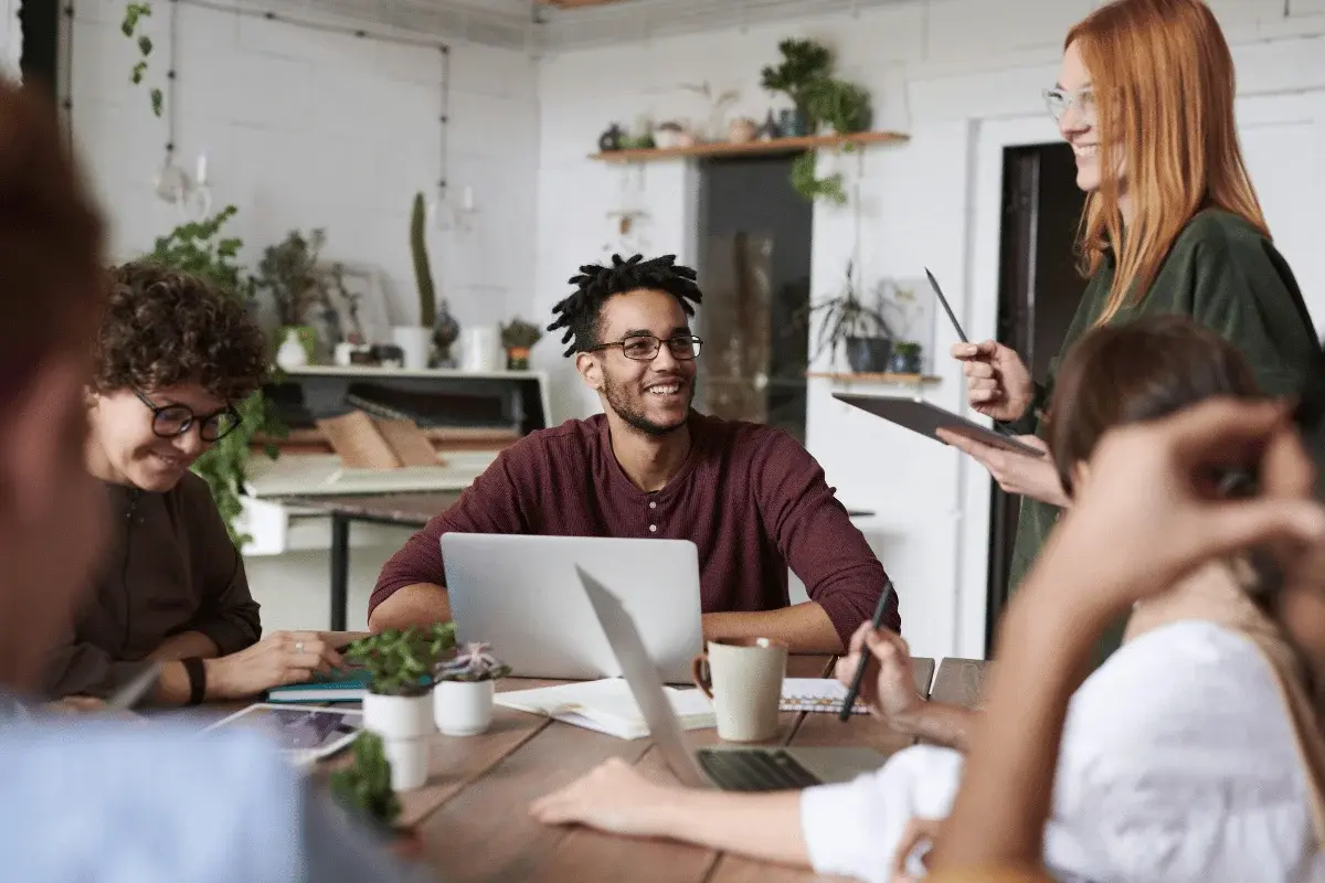 A group of people in a cozy office, engaged in a meeting. One person stands, smiling, holding a tablet, while others sit, using laptops and chatting.