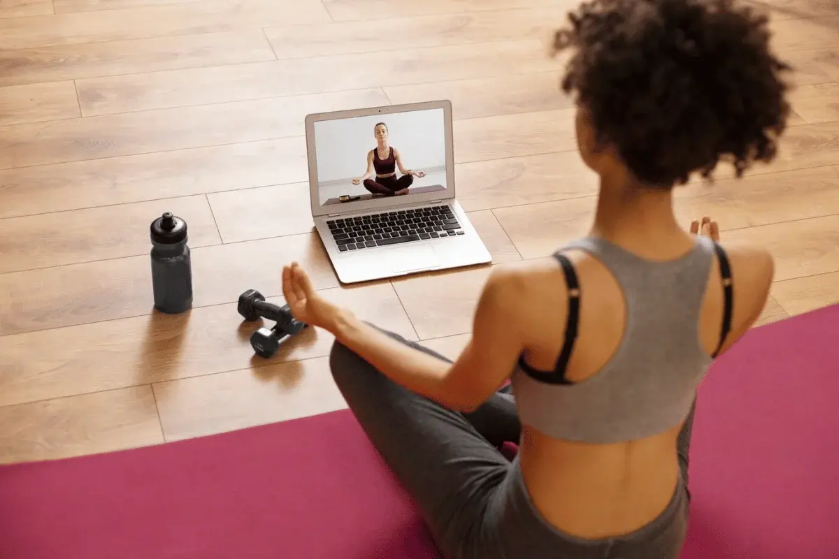 A person sits cross-legged on a yoga mat, following an online meditation class displayed on a laptop, with dumbbells and a water bottle nearby.